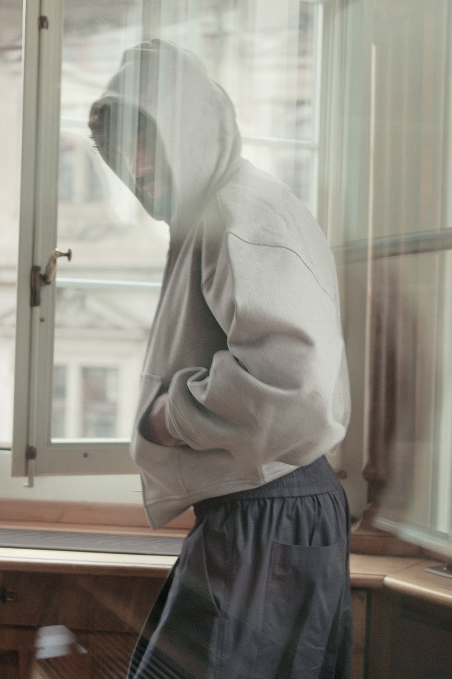 A person wearing a light gray oversized hoodie and dark gray sweatpants stands by a large window in a room with wood trim, looking out.
