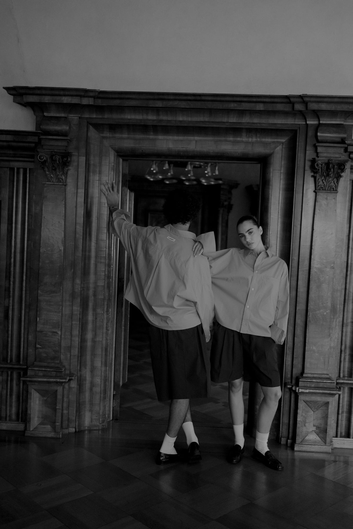 A black and white photograph of two models in a grand, historic interior with an ornate wooden doorway. Both are wearing oversized light-colored button-up shirts and dark shorts, with one model facing away and the other facing forward.