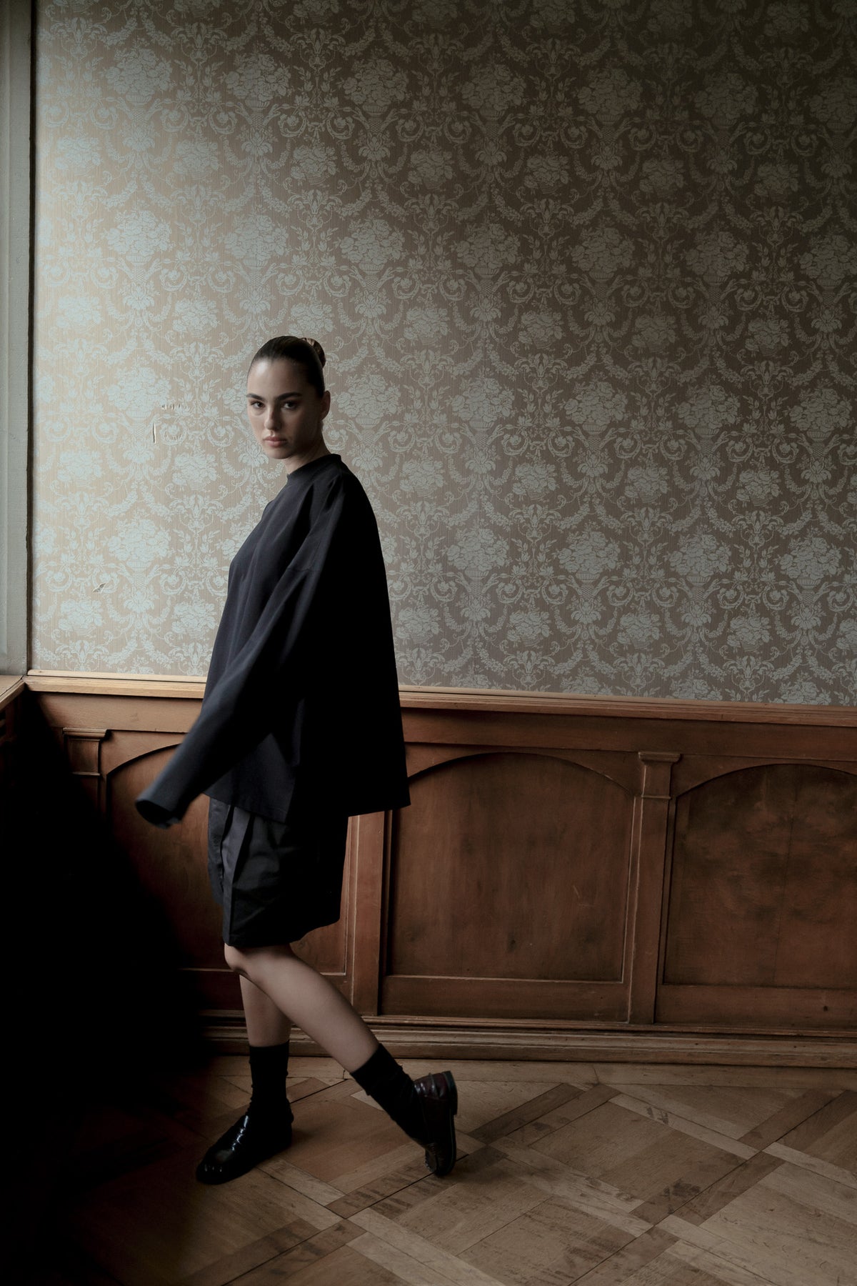 A model walks across a wooden parquet floor against a wall with ornate damask wallpaper and wooden paneling, wearing a loose oversized black jacket, black shorts, black socks, and black leather shoes.