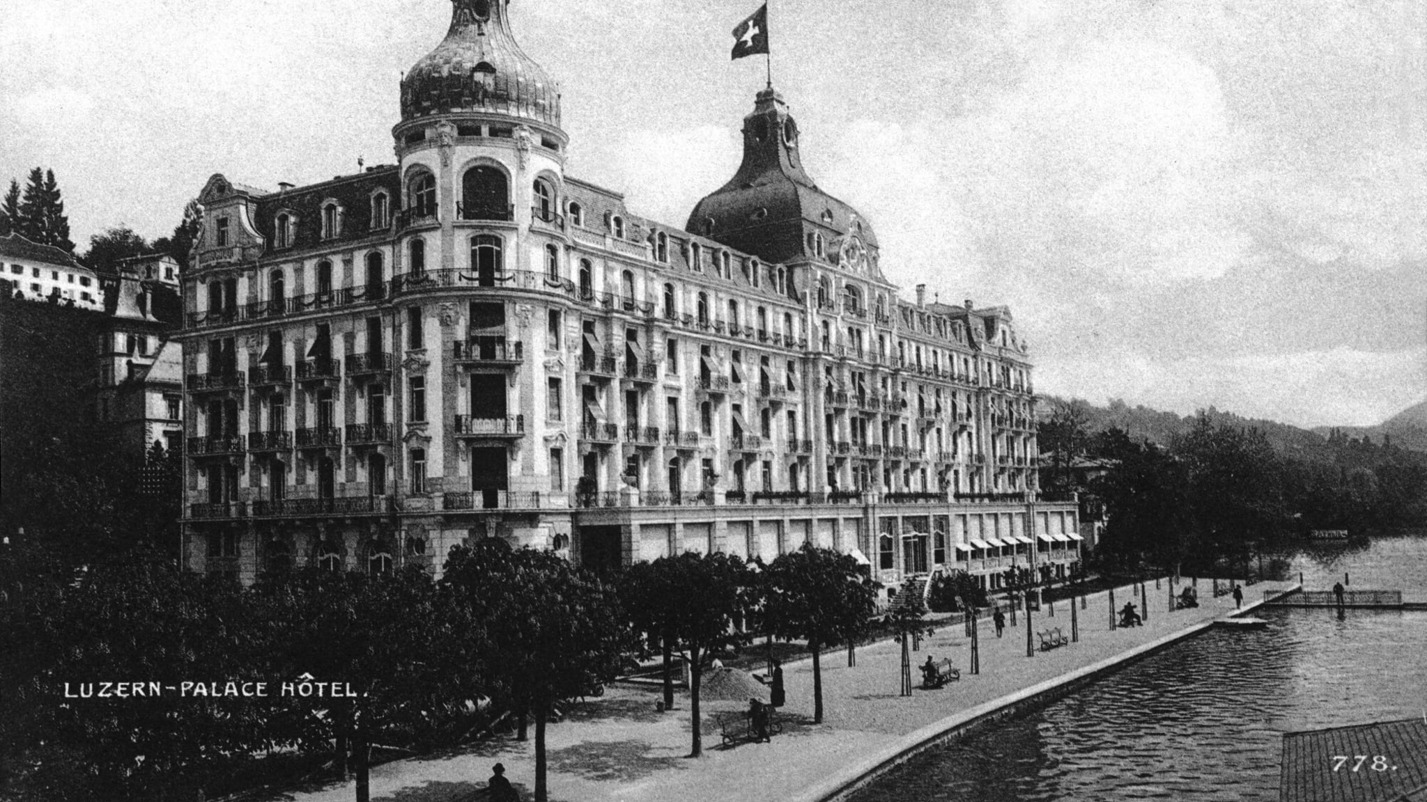 Historic landscape view of Palace Hotel Luzern on the shore of Lake Lucerne, with the Alps in the distance.
