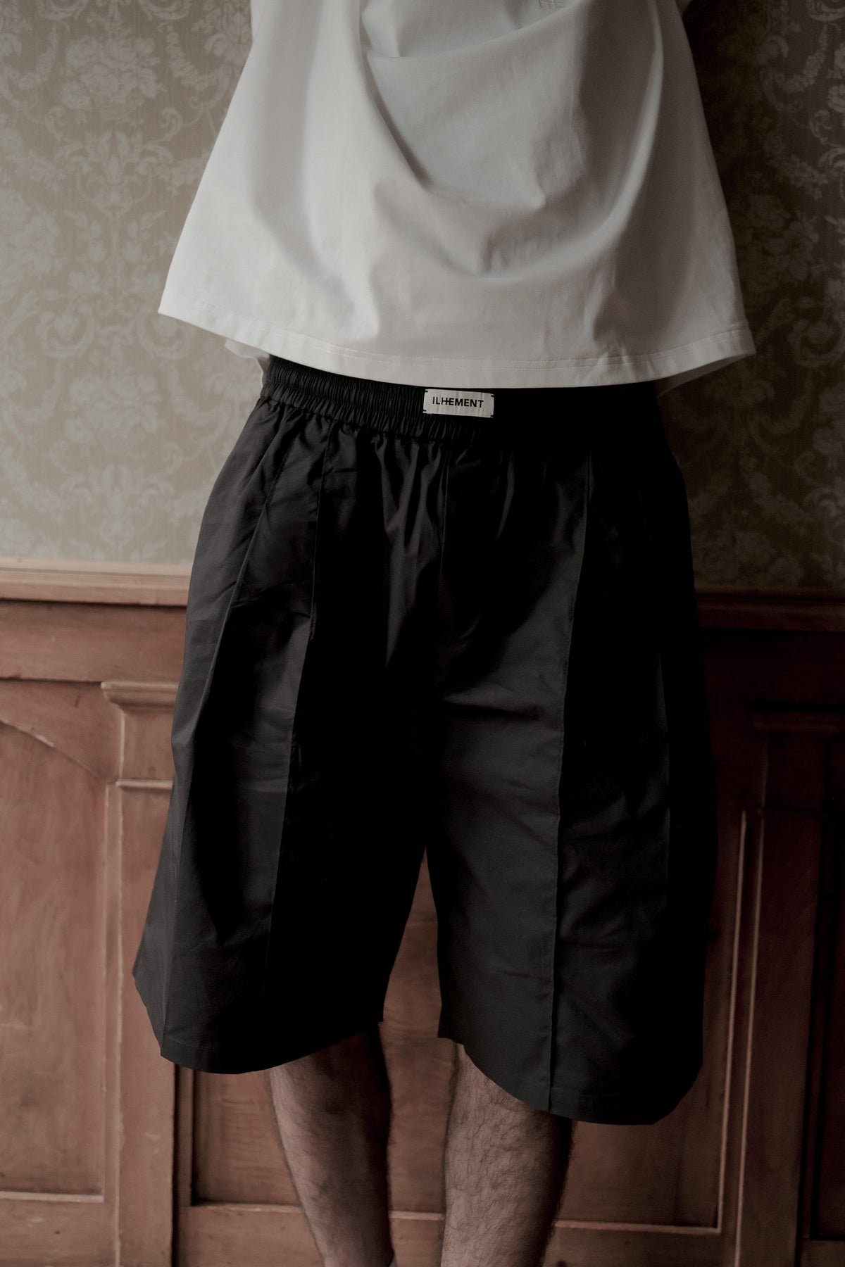 A person wearing black pleated shorts and a white t-shirt, standing in front of a wooden panel wall and patterned wallpaper. The brand name 'LYRANT' is visible on a small white label at the waistband.