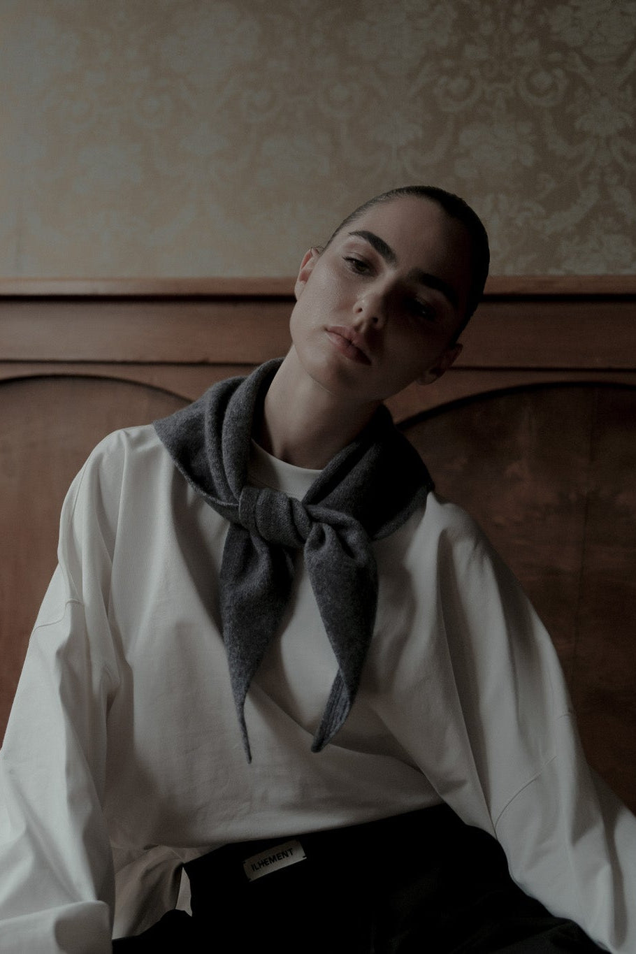 A person with a sleek hair back look wearing a white shirt and a dark gray knitted triangle scarf, seated in front of a dark wood-paneled wall.