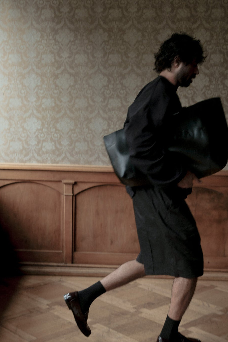 A man in a black outfit carrying a large black leather bag walks through a dimly lit room with patterned wallpaper and wood paneling.