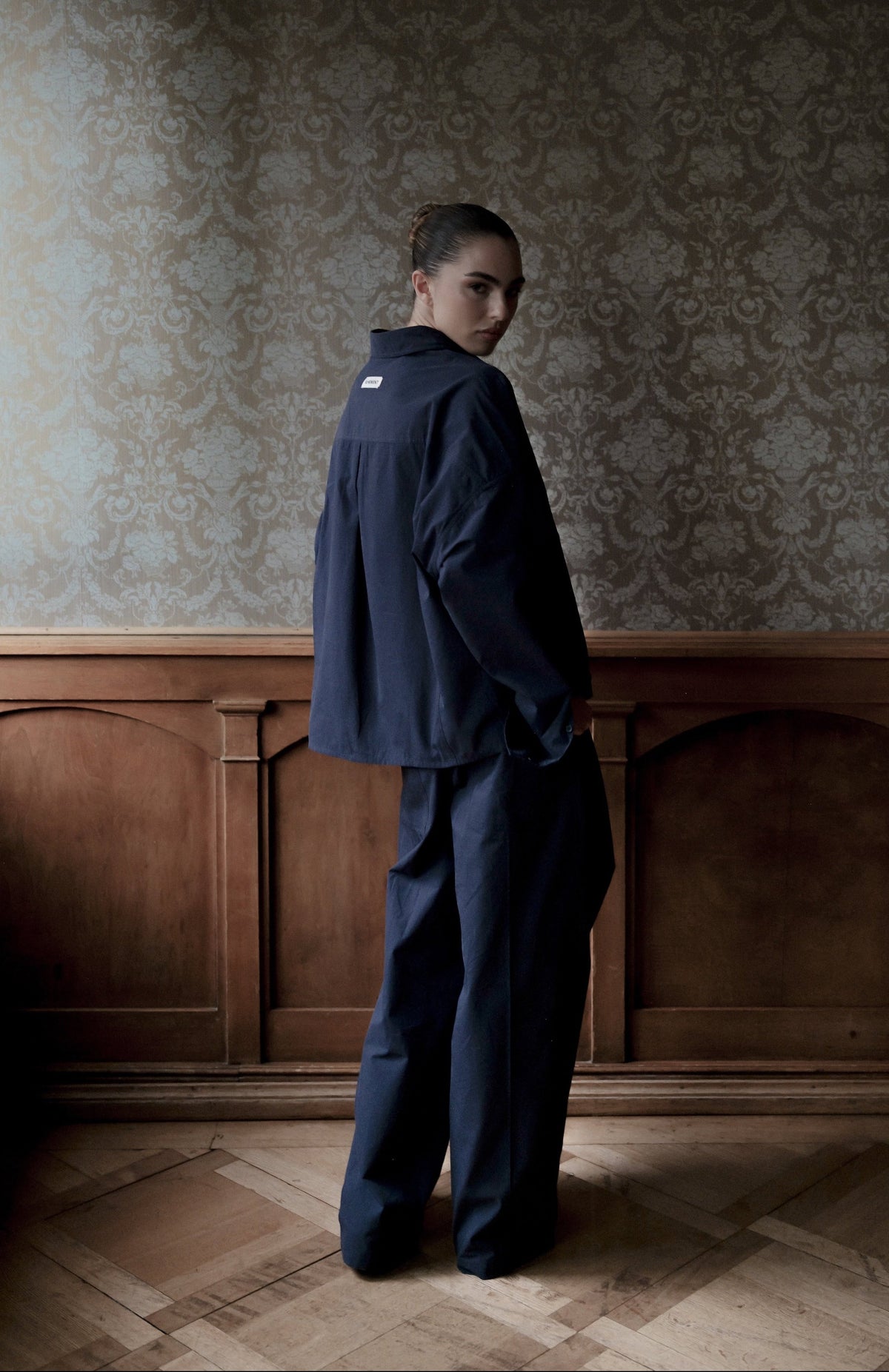 A model stands in profile wearing a dark navy blue jacket and matching trousers in a room with ornate wallpaper and wood paneling.