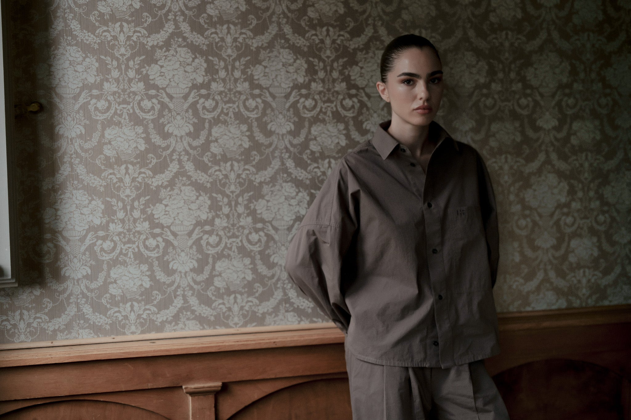 A model stands in profile wearing a Taupe jacket and matching trousers in a room with ornate wallpaper and wood paneling.