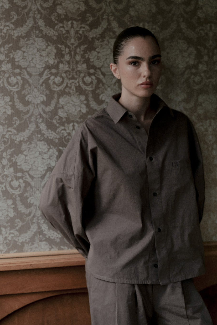 A model stands in profile wearing a Taupe jacket and matching trousers in a room with ornate wallpaper and wood paneling.