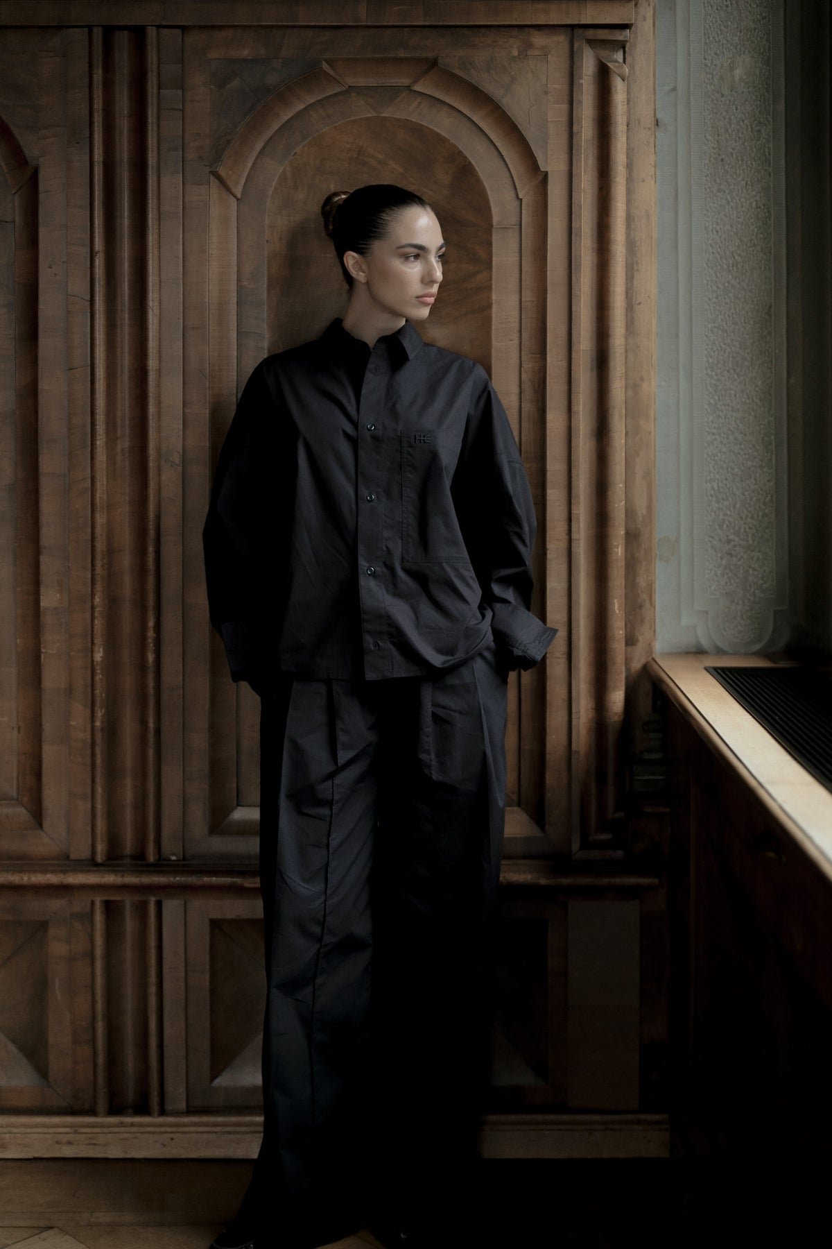 A model stands in a dimly lit interior with dark wood paneling, wearing a loose black long-sleeve button-front top and matching wide-leg trousers.