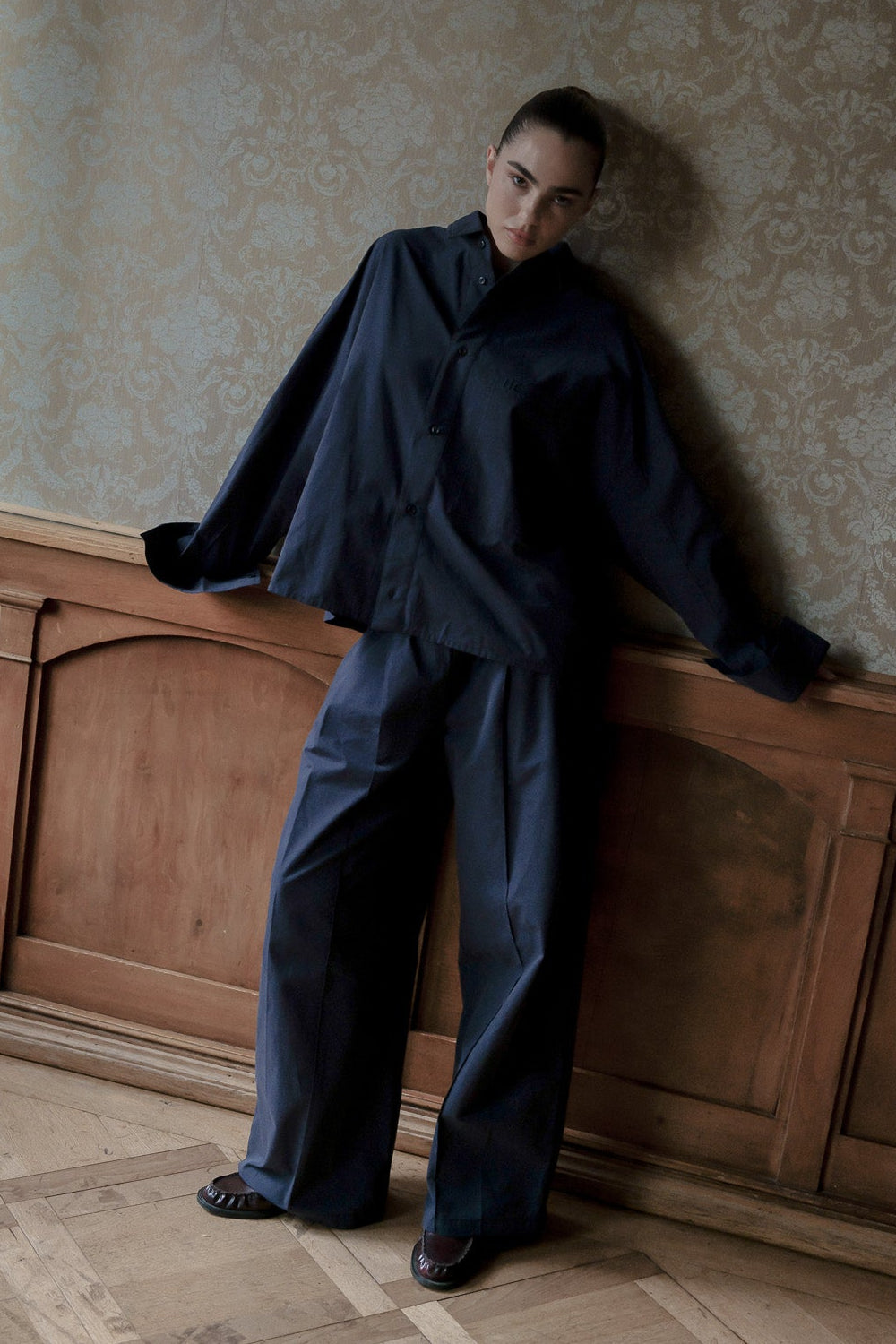 A model wearing a dark navy blue oversized long-sleeve shirt and matching wide-leg trousers, leaning against a wooden panel wall in an interior room with patterned wallpaper.