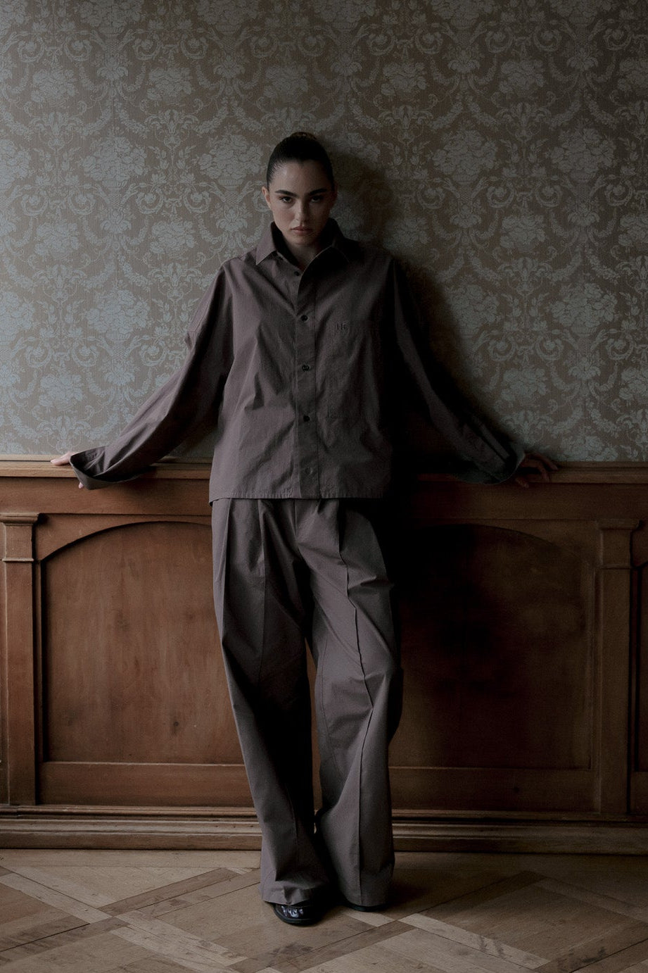 A model stands in a dimly lit room with patterned wallpaper and a wooden wainscoted wall, wearing a matching dark brown long-sleeve button-up shirt and wide-leg trousers.