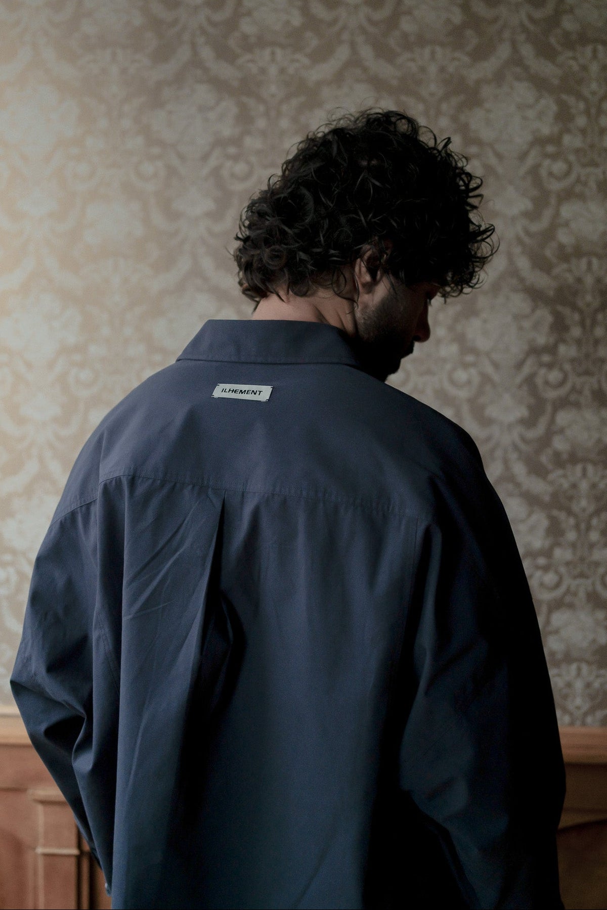A man with curly dark hair is shown from behind wearing a dark navy blue overshirt with a small white 'STAMPEY' brand label on the back collar, set against a patterned beige wallpaper background.