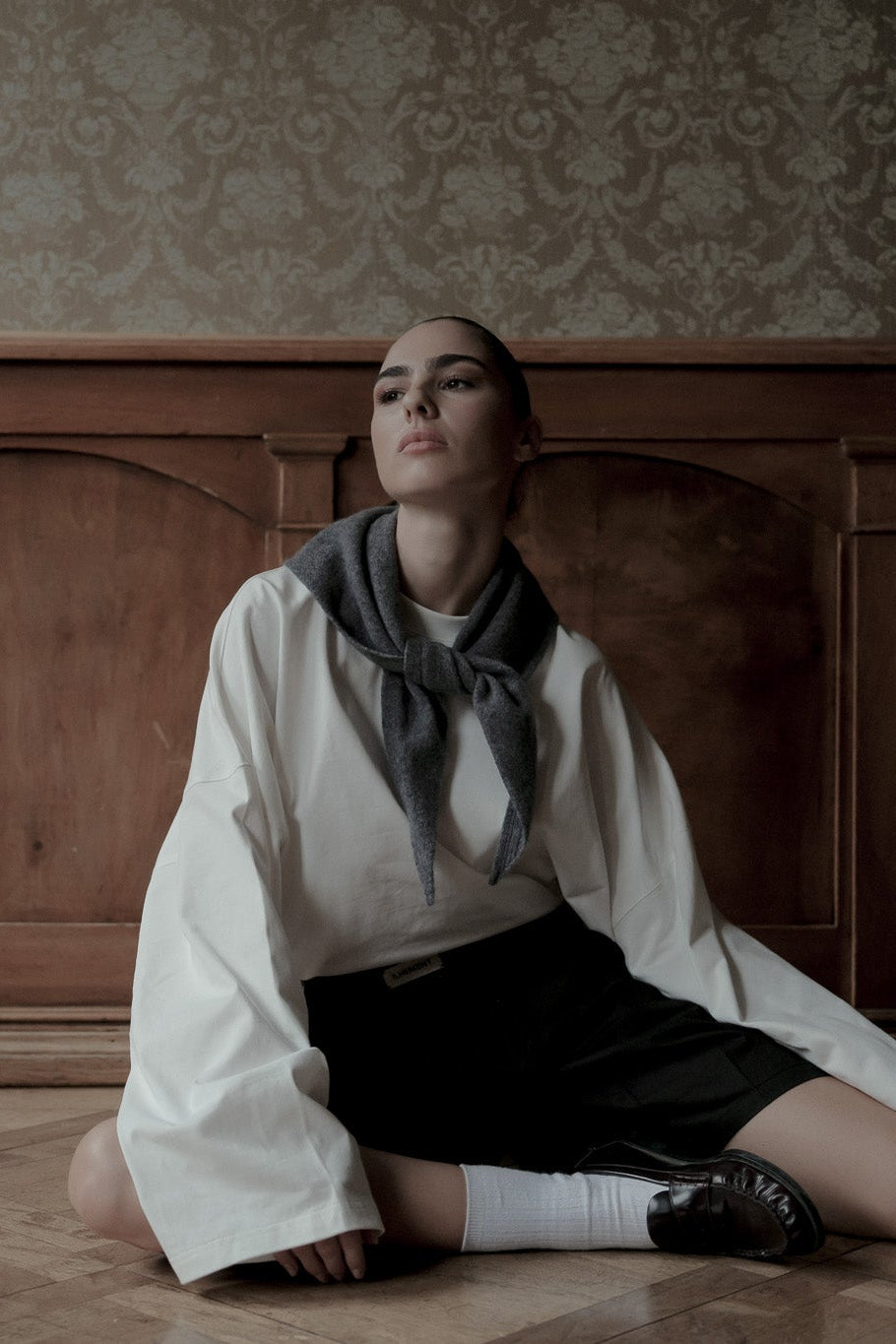 A model sits cross-legged on a wooden floor against a dark wood paneled wall and patterned wallpaper, wearing a white shirt with a dark gray scarf, a black skirt, white socks, and black shoes.